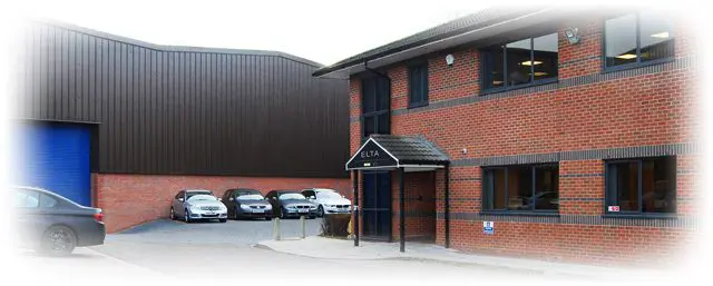 A brick office building with a sign above the entrance that reads "ELTA." Several cars are parked in a lot adjacent to the building. The building has large windows and is next to a larger, dark-colored warehouse.