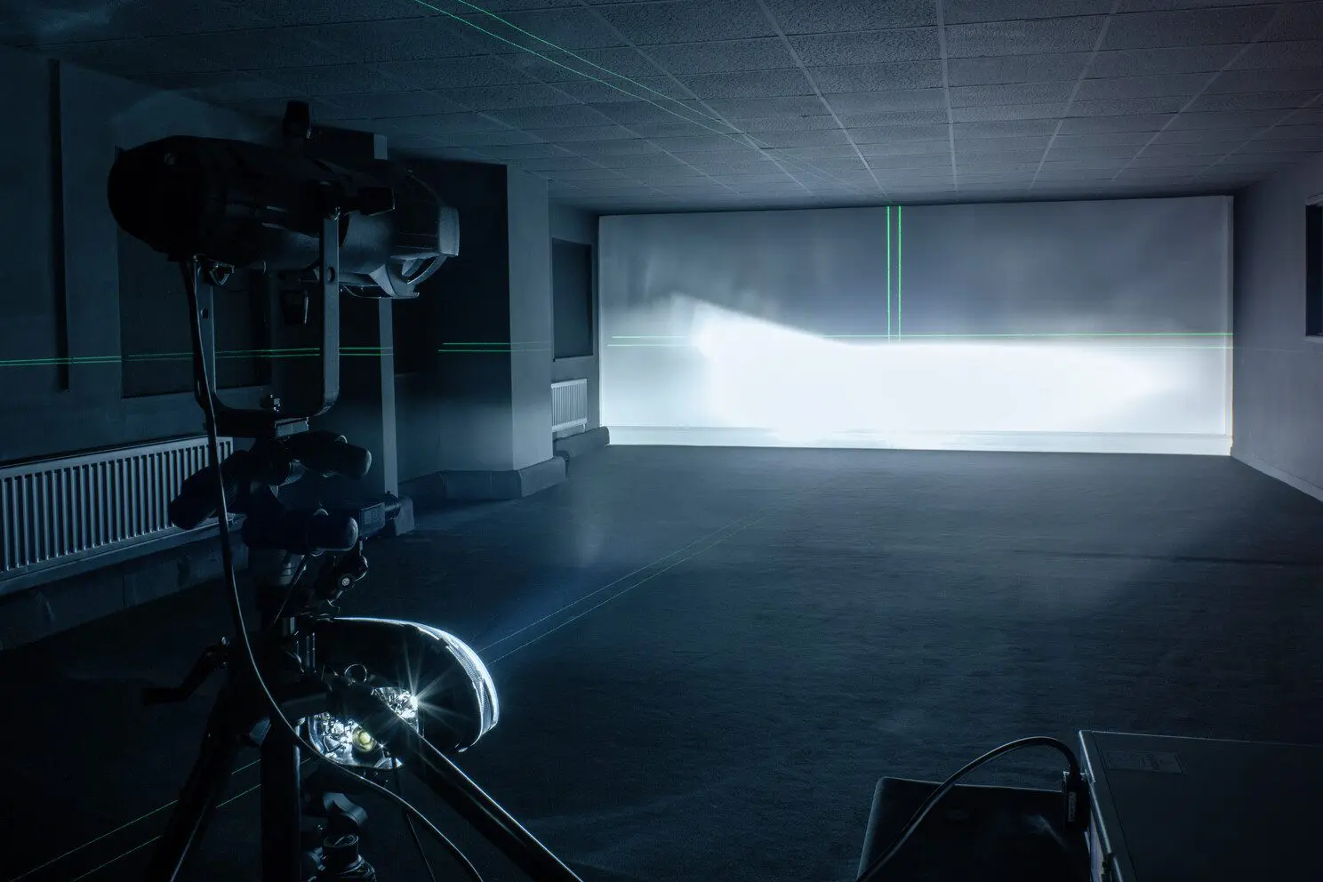 A dark room with a large white screen at the far end, green laser lines forming vertical and horizontal patterns on the screen, and a camera or projector mounted on a tripod in the foreground.