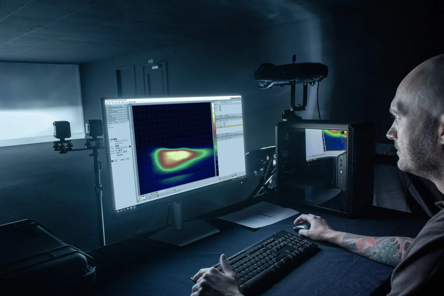 A person working at a computer station in a dark room, focused on a screen displaying a colorful heat map or simulation graph, with additional monitors and equipment in the background.