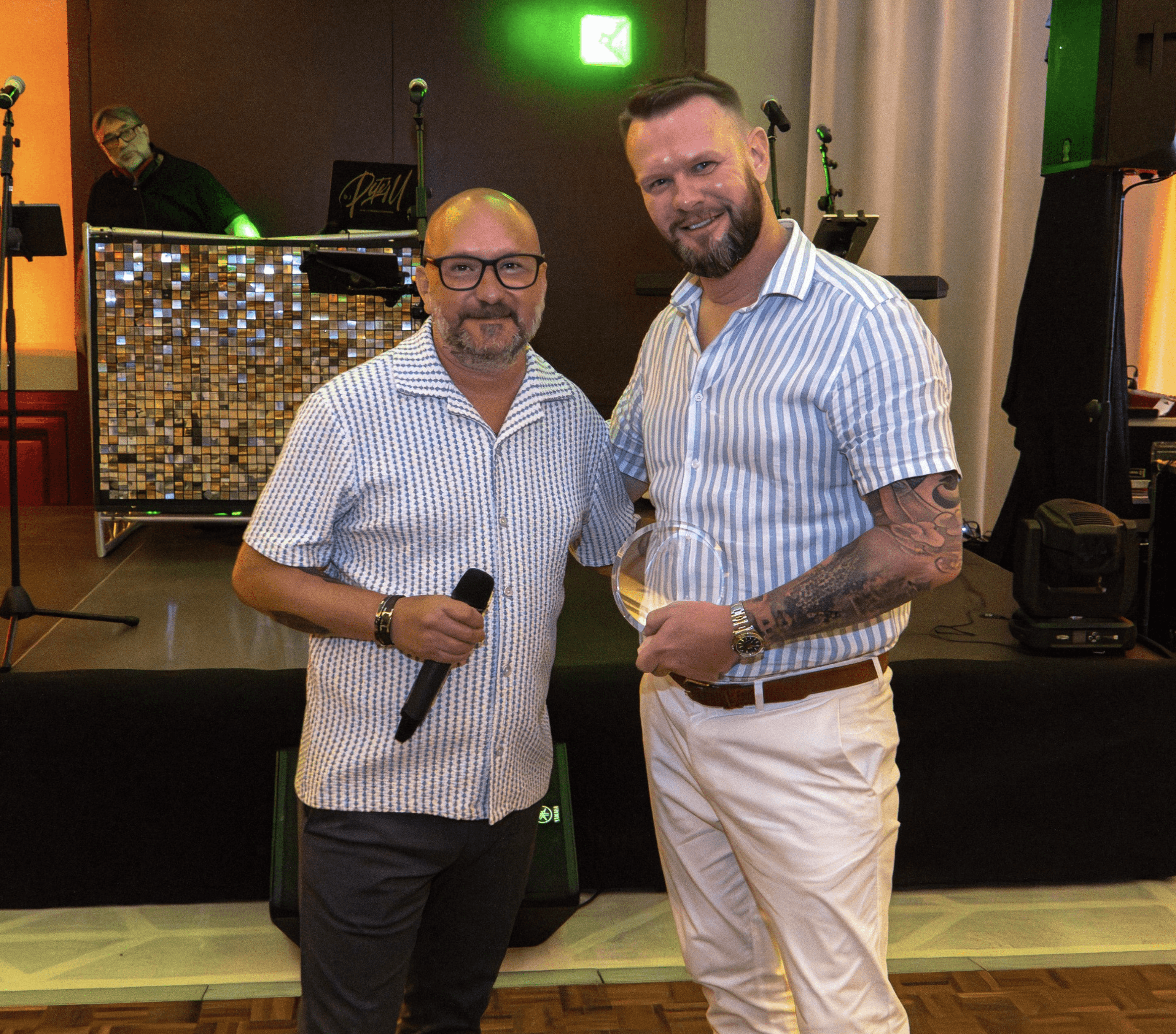Two individuals wearing striped shirts stand together in front of a stage with musical equipment and a DJ. One person holds a microphone, and the other has a tattooed arm