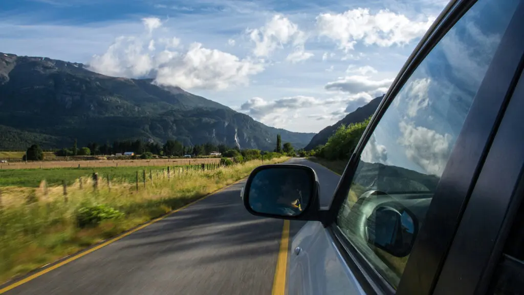 A view from a vehicle driving along a rural road, showing mountains, fields, and clouds reflected in the vehicle’s side window and mirror.