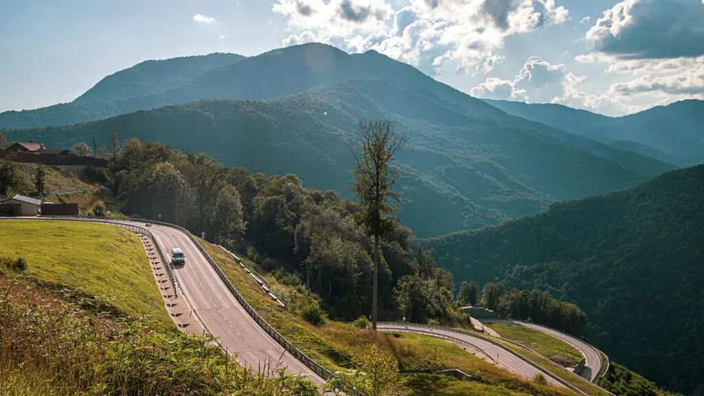 A winding mountain road with a vehicle traveling along it, surrounded by forested hills and large mountains under a partly cloudy sky.