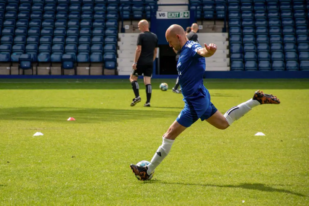 A football player striking a ball on a grass pitch during training, with other players and empty stadium seating in the background.