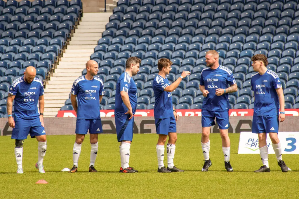 The image shows a team gathered on a football pitch inside a stadium, wearing matching blue kits.