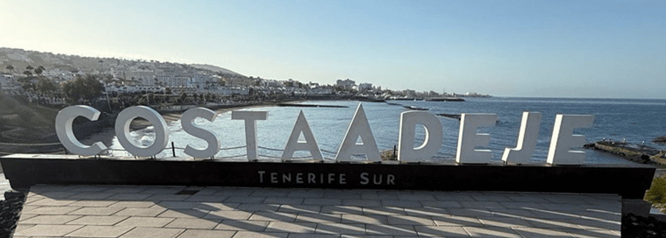A large sign reading "COSTA ADEJE" with smaller text "TENERIFE SUR" below it, located on a promenade overlooking the sea. A coastal town and the ocean are visible in the background.