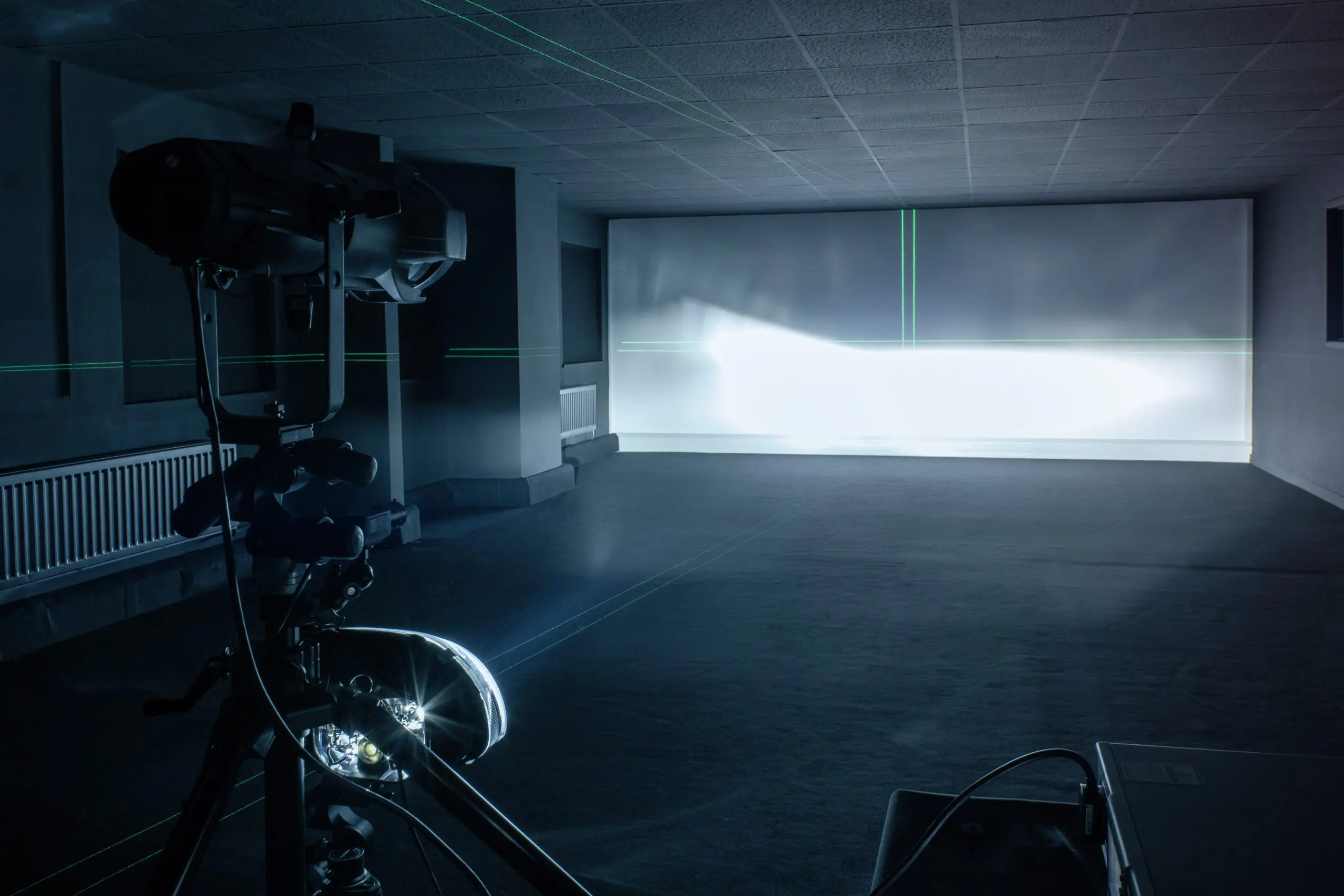 A headlamp testing setup projecting a beam pattern onto a wall in a dark room, with alignment laser lines visible.